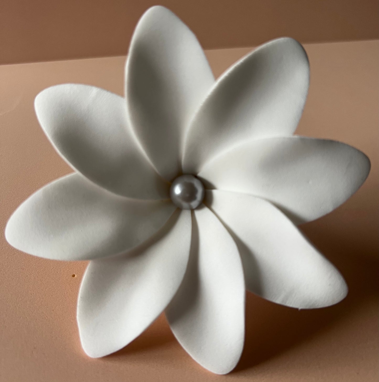 Fleur artificielle blanche composée de deux rangées de cinq pétales arrondis, avec une perle nacrée au centre. Elle repose sur un fond rose poudré, créant une ambiance douce et élégante.