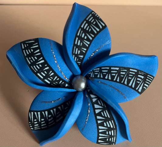 Fleur artisanale à cinq pétales en mousse bleue vive, chaque pétale étant décoré d’un motif géométrique noir et blanc évoquant des motifs ethniques. Les pétales sont soulignés de fines paillettes argentées. Au centre de la fleur, une perle nacrée gris anthracite apporte une finition élégante et brillante.