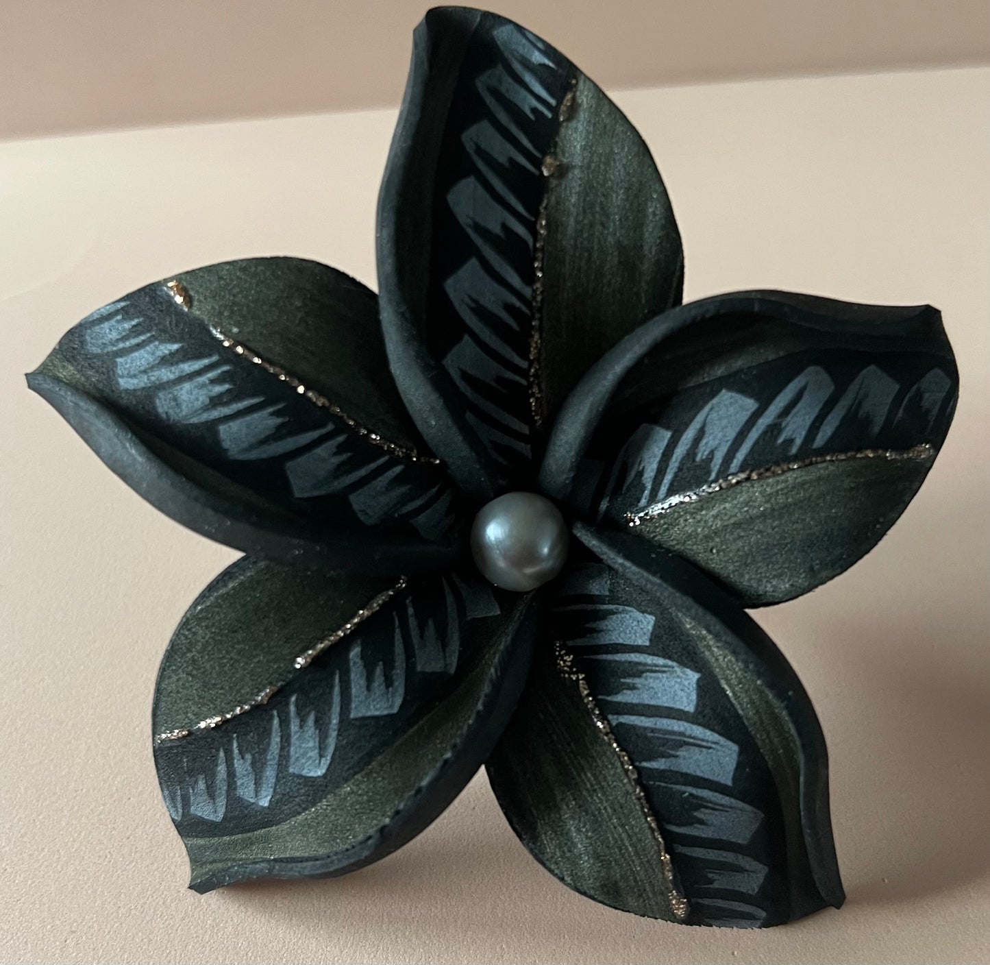 Fleur artificielle noire à cinq pétales ornés de motifs gris et de fines lignes dorées scintillantes. Une perle sombre est placée au centre, sur un fond beige rosé.