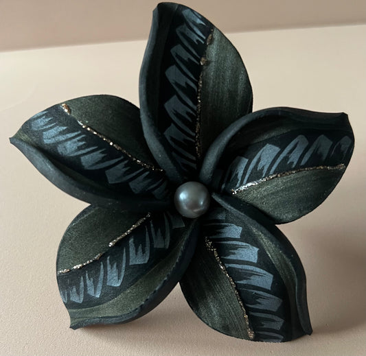 Fleur artificielle noire à cinq pétales ornés de motifs gris et de fines lignes dorées scintillantes. Une perle sombre est placée au centre, sur un fond beige rosé.