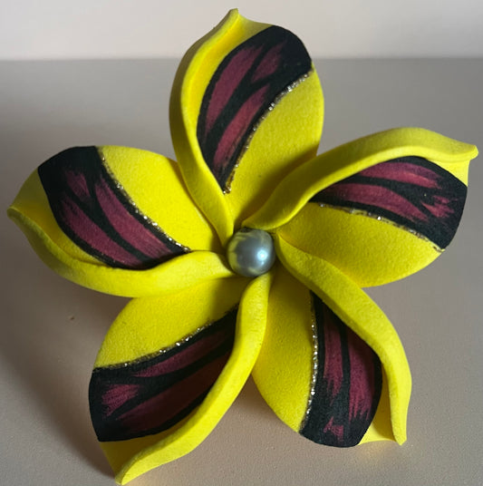 Fleur décorative à cinq pétales réalisée en mousse jaune, ornée de motifs noirs et bordeaux sur chaque pétale, avec des touches de paillettes argentées. Le centre de la fleur est agrémenté d'une perle gris nacré. La fleur est posée sur une surface beige claire.